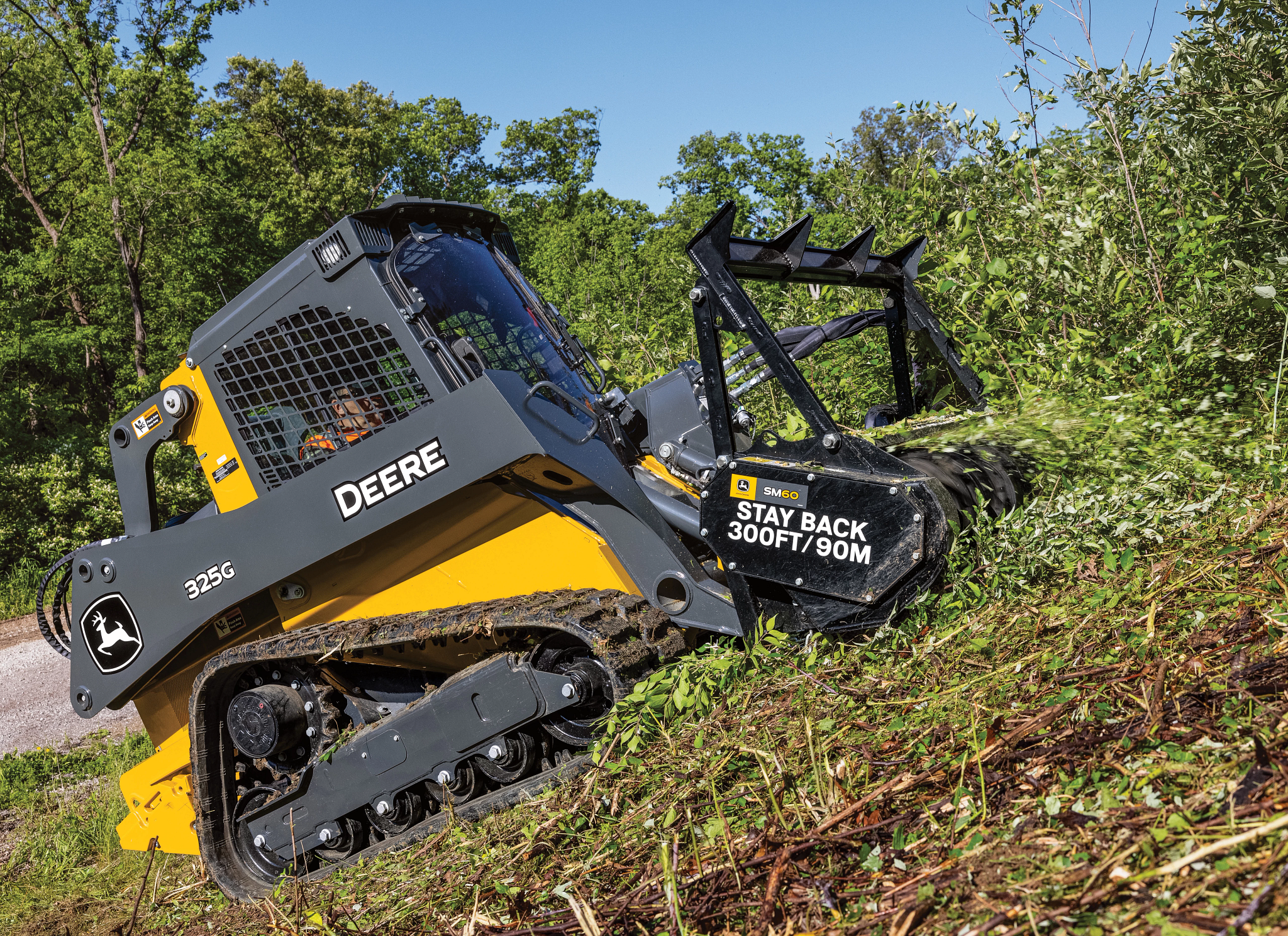 A tracked compact loader on a hillside using a front-mounted mulching head to clear dense vegetation, sending cut material forward