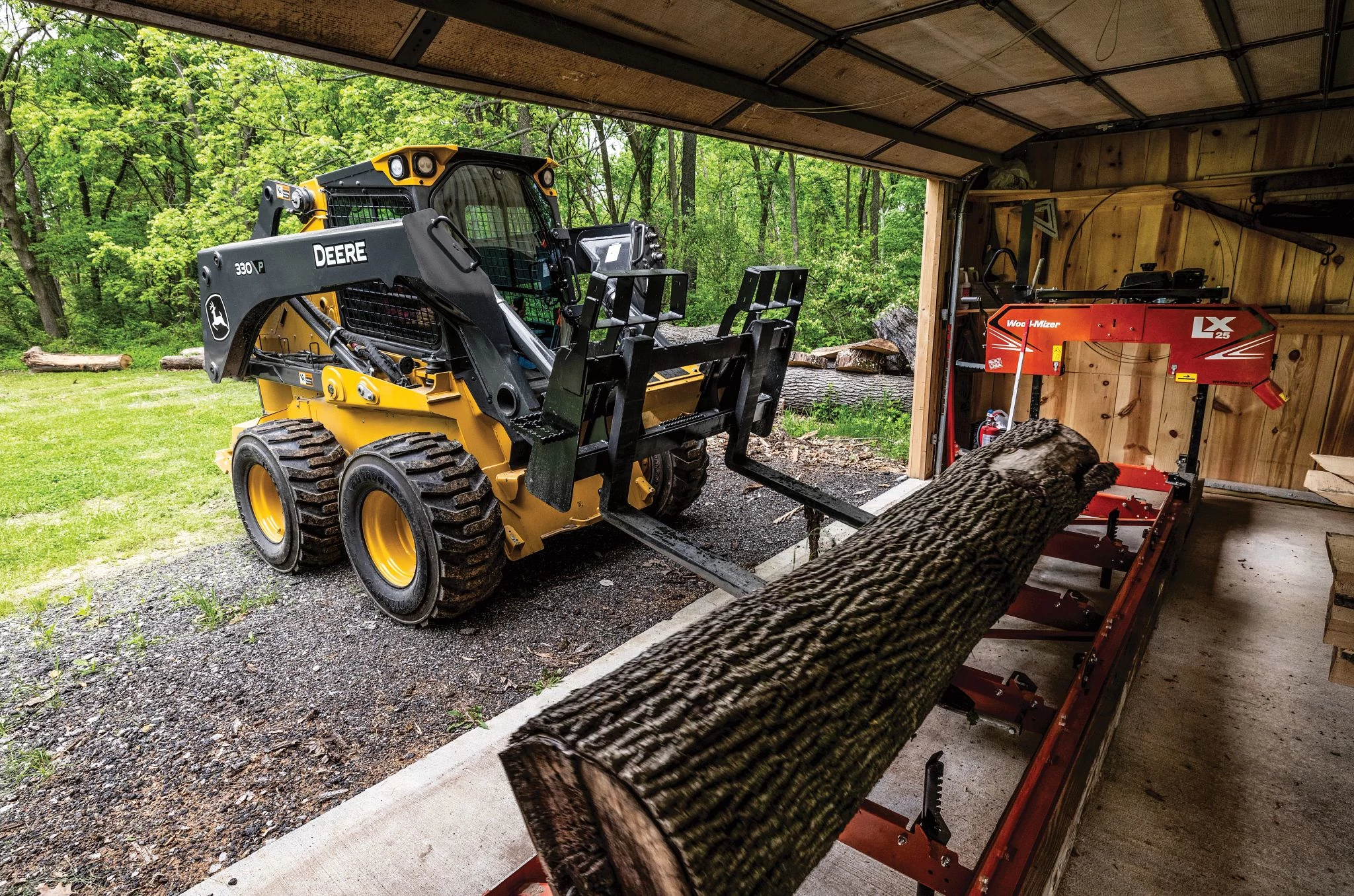 A yellow John Deere skid steer loader operates inside an open-sided shed, using pallet forks to lift and position a large cut log onto a red portable sawmill.