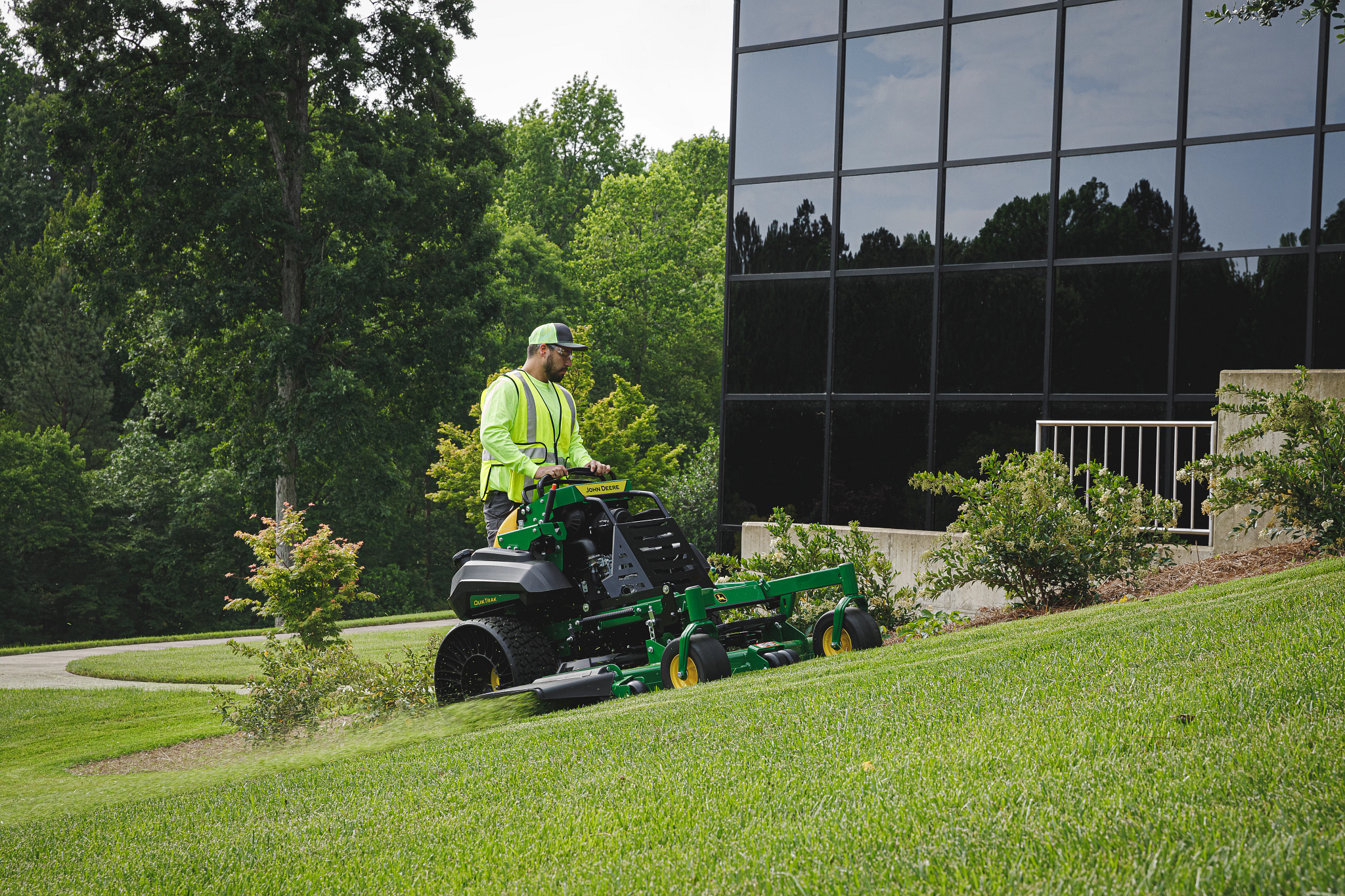 Yellow-vested operator mowing grass on an incline in front of a black glass building on a Q850R Stand-on QuikTrak mower with 60-in. 7Iron PRO deck