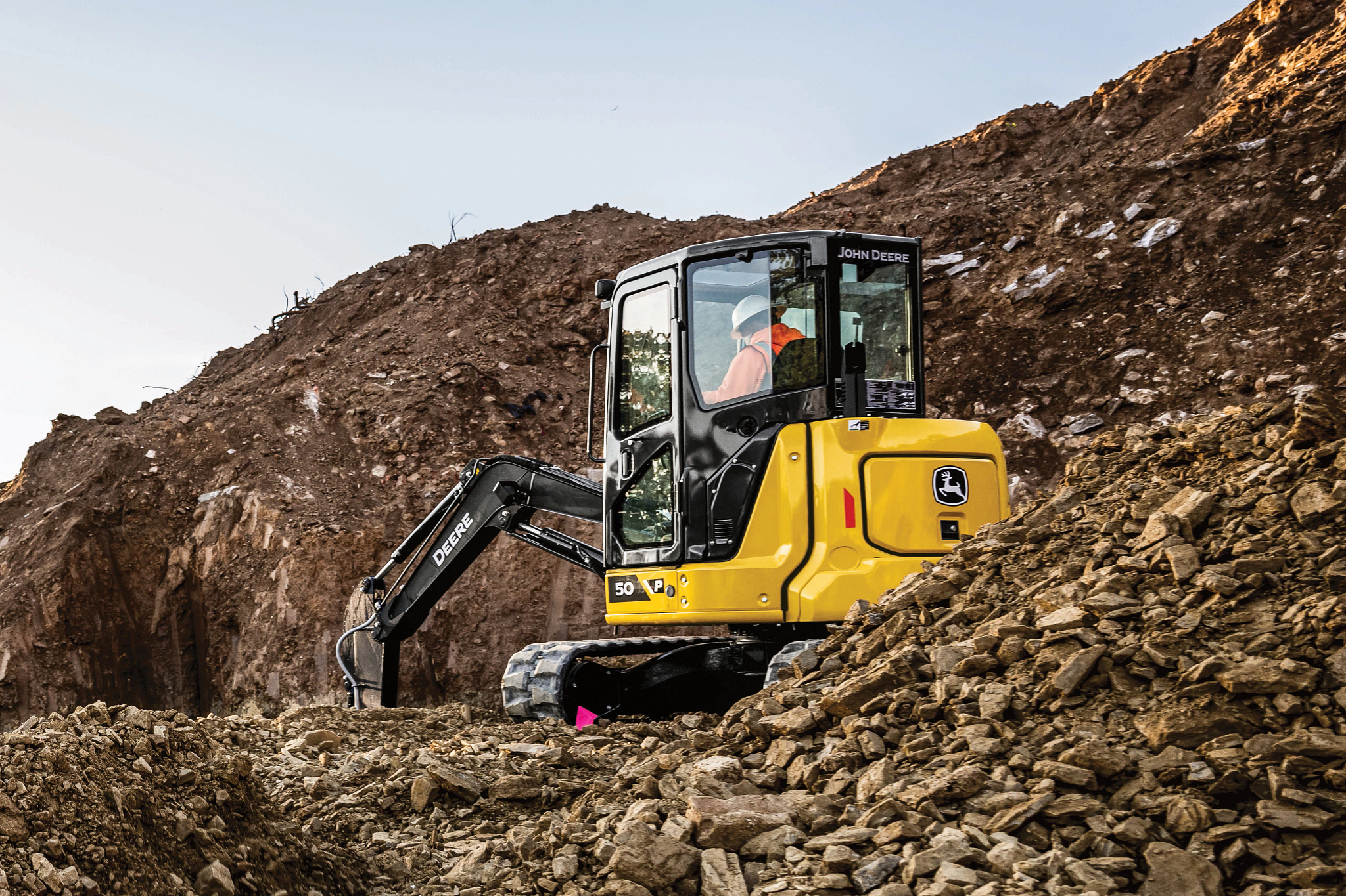 Compact excavator operating on a rocky slope at a construction site, with the machine’s arm digging into the dirt and a large mound of earth rising behind it