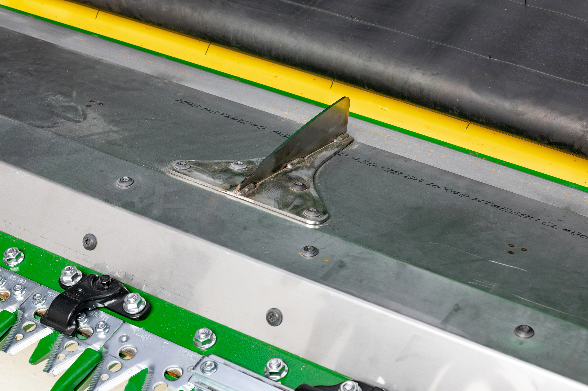 A close-up of a metal crop guide mounted on a green and silver combine draper, with cutting components and a belt visible around it