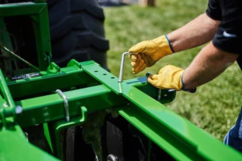 Close-up of  an L-shaped pin being adjusted on a green agricultural implement frame, with metal brackets, fasteners, and a tractor tire.