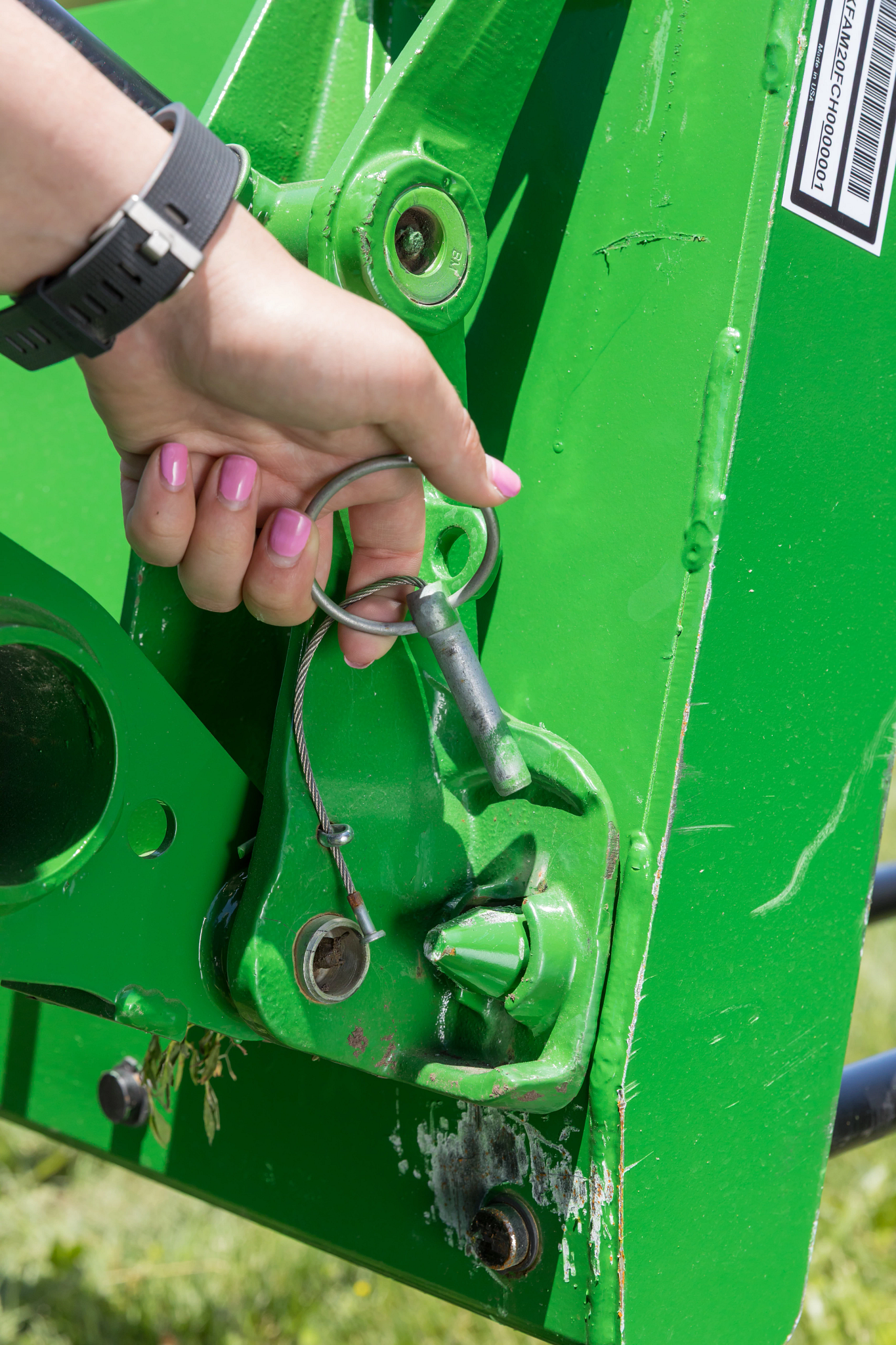 Close-up of a metal safety pin with cable being inserted into a green agricultural attachment’s mounting bracket, showing the locking mechanism, bolts, and painted steel components.