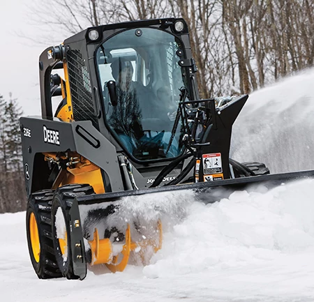 A compact loader equipped with a front snow blower clearing a path by blowing snow forward and to the side, with leafless trees in the background