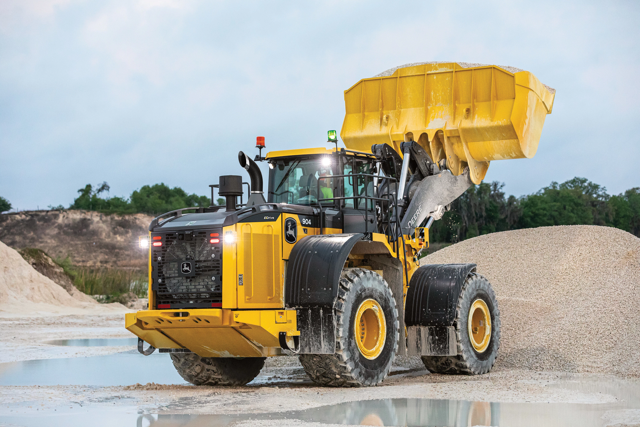 Rear right view of a 904 X-Tier Wheel Loader in a watery gravel quarry with fully-loaded bucket suspended in the air above a gravel pile.