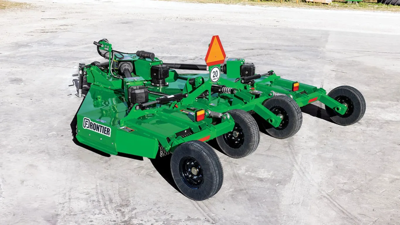Green multi-spindle rotary mower attachment with four tires, a slow-moving vehicle triangle, and multiple cutting decks, parked on a concrete surface in an outdoor setting