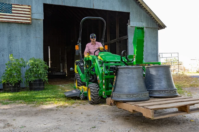 Green compact tractor carrying two large metal bells on a front loader pallet.
