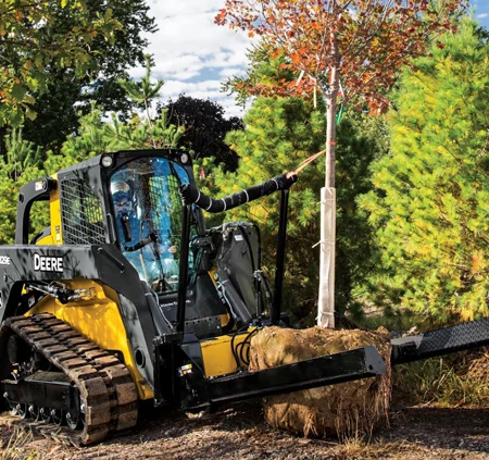 Front right view of a track loader carrying a tree and its root bundle in a wooded area with an NF44 nursery fork attachment