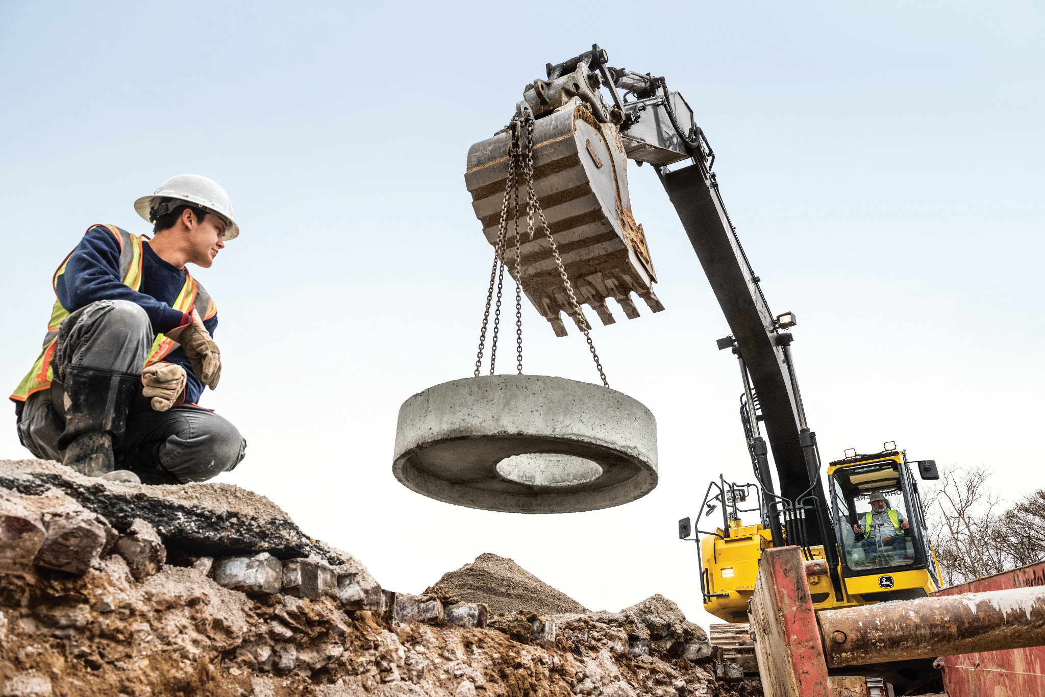 Construction worker kneeling on rocky ground while a yellow excavator with a grapple bucket lifts a large circular concrete ring using chains