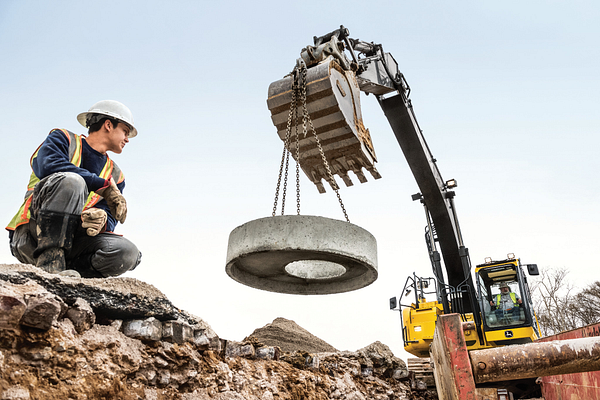 Construction worker kneeling on rocky ground while a yellow excavator with a grapple bucket lifts a large circular concrete ring using chains
