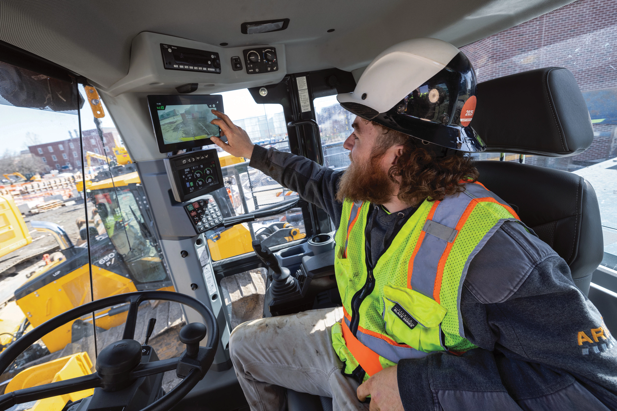Interior view of a heavy construction vehicle cab showing an operator seated at the controls, wearing a hard hat and a high-visibility safety vest