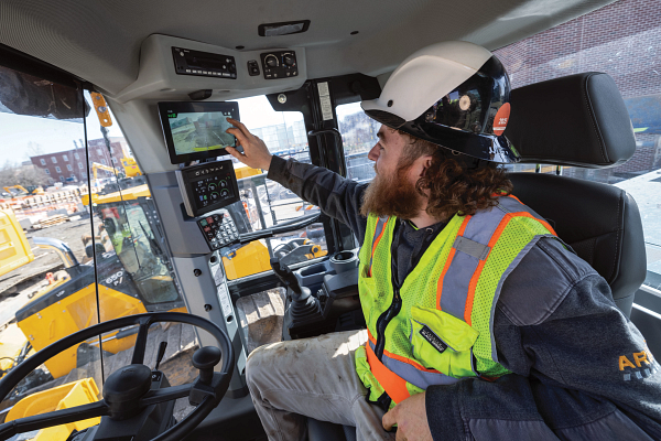 Interior view of a heavy construction vehicle cab showing an operator seated at the controls, wearing a hard hat and a high-visibility safety vest