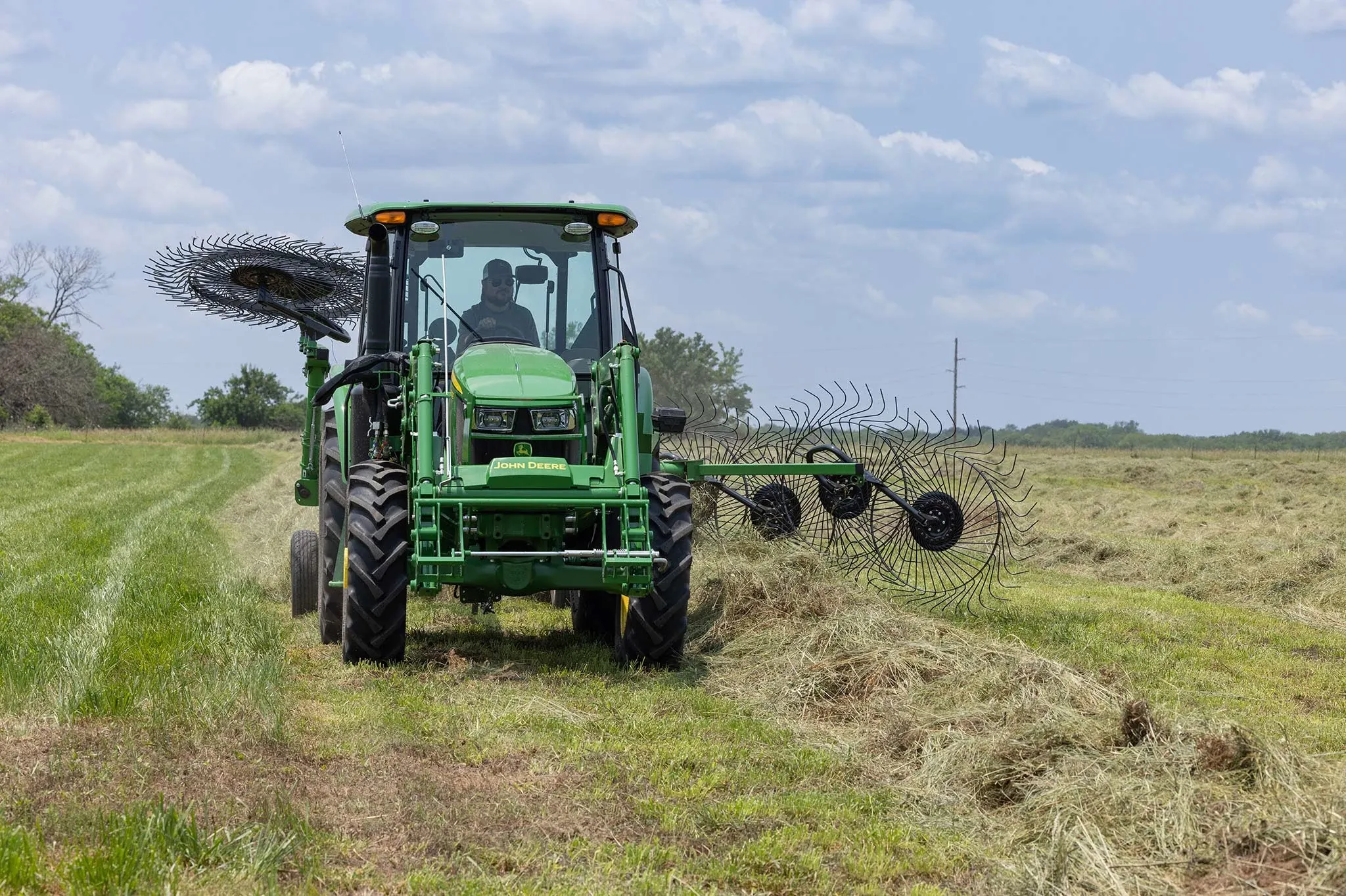 Front view of a 5E series tractor driving along a hay windrow with a WR3212 wheel rake at work behind it