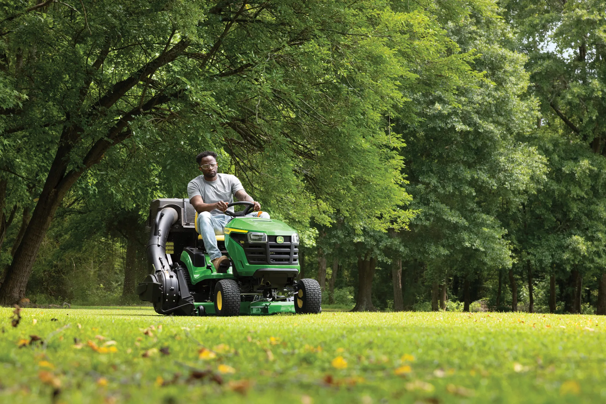 Front right view of an operator using an S220 Lawn Tractor with attached material collection system on a wooded lot