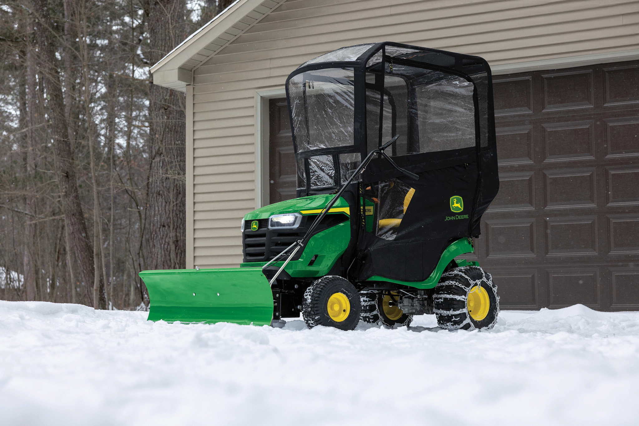 Front left view of an S140 Lawn Tractor with black weather enclosure parked on a snowy driveway in front of a closed garage.