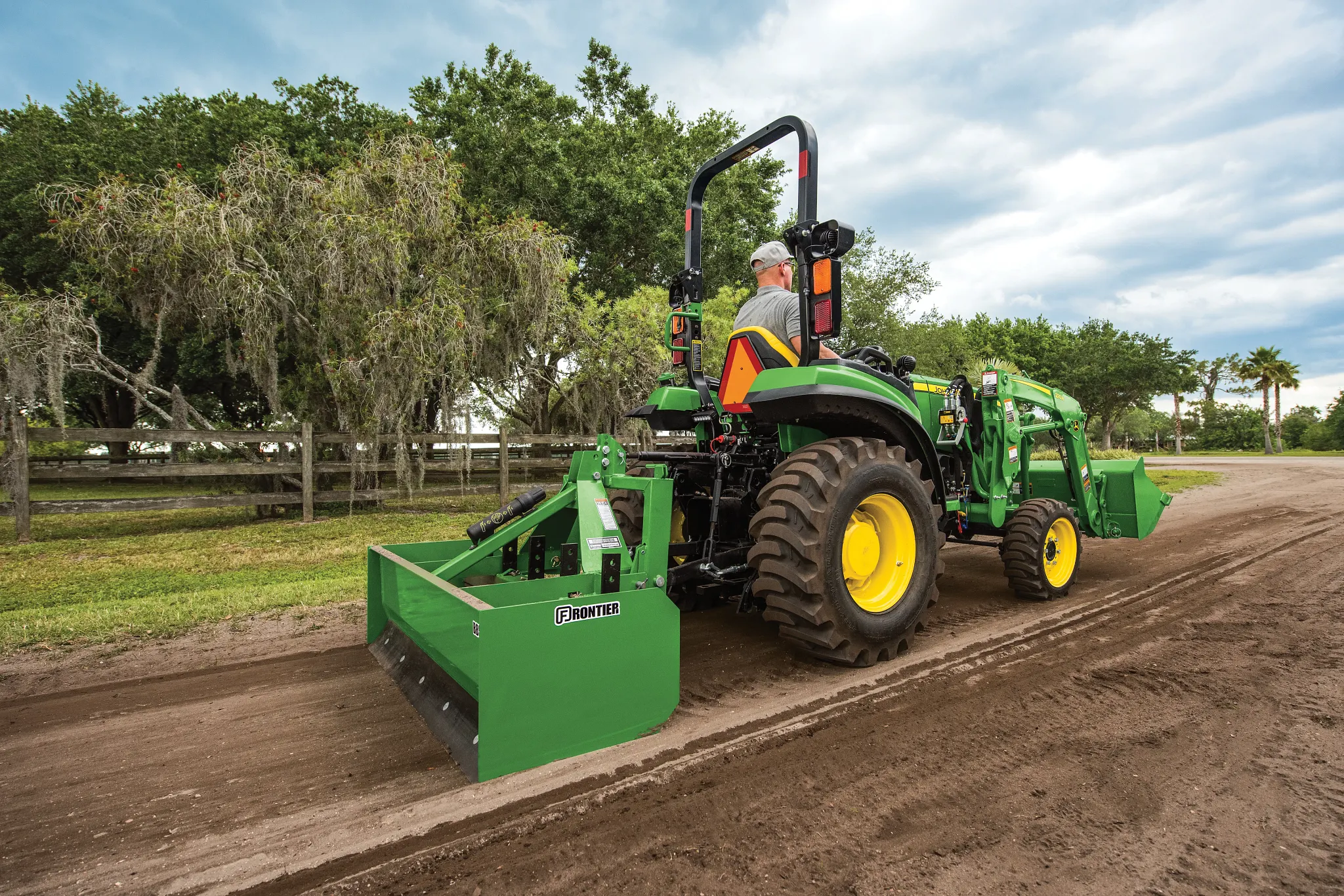 Green compact tractor with large rear tires and a rear grading box drives along a dirt path in a rural setting, bordered by grass, trees, and a wooden fence under a cloudy sky