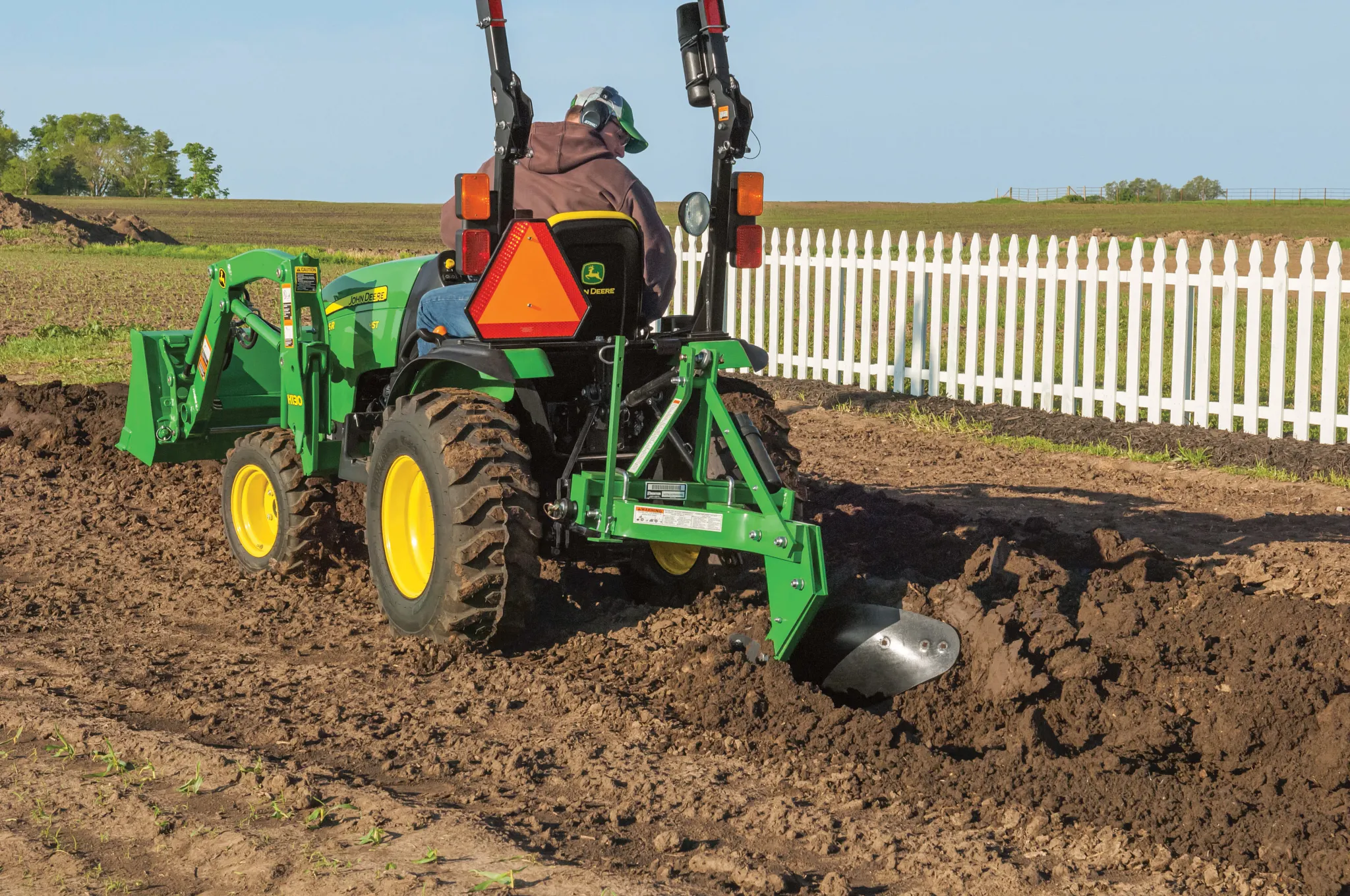 Green tractor with a rear plow attachment cuts a trench through freshly tilled soil near a white picket fence, with a driver seated at the controls and open farmland in the background.