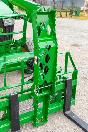 Close-up of a front loader attachment with a vertical grapple or upright frame featuring cutout holes, mounted above pallet forks.