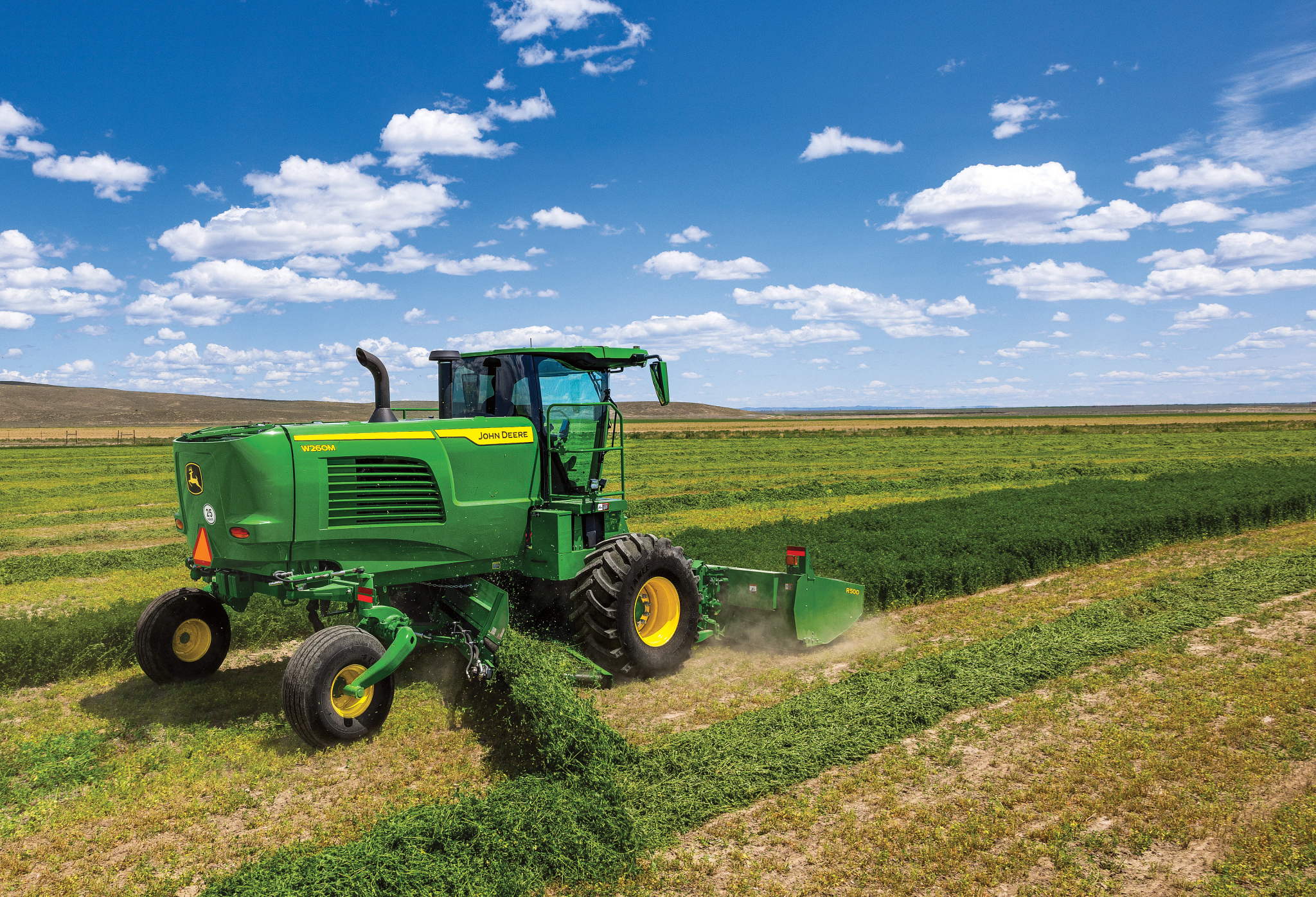 John Deere self‑propelled windrower cutting hay in an open field, side view showing mower‑conditioner forming a windrow behind the machine under a blue sky with scattered clouds.