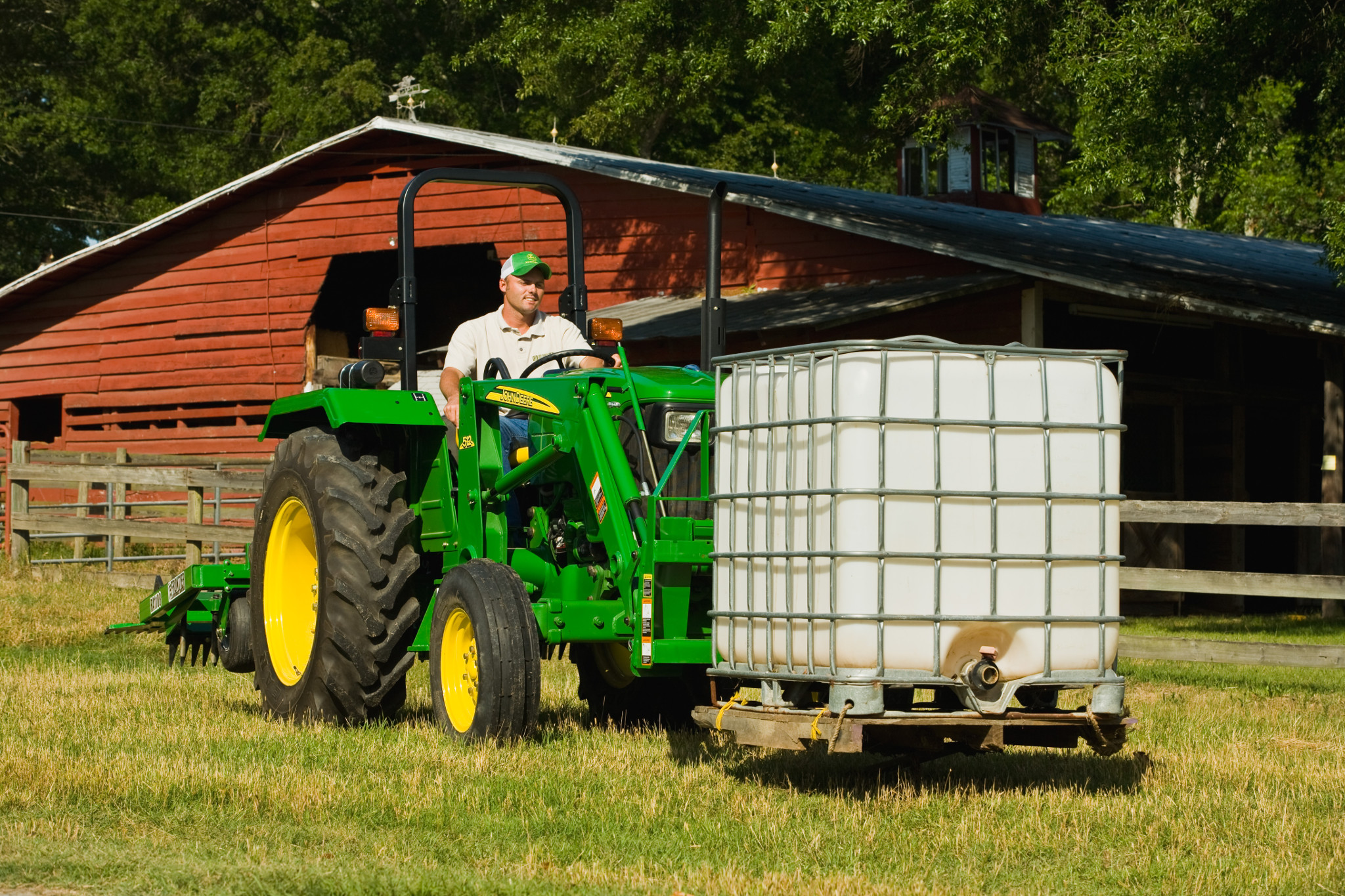A man operating a tractor with a pallet fork transporting a large square water tank in front of a red barn