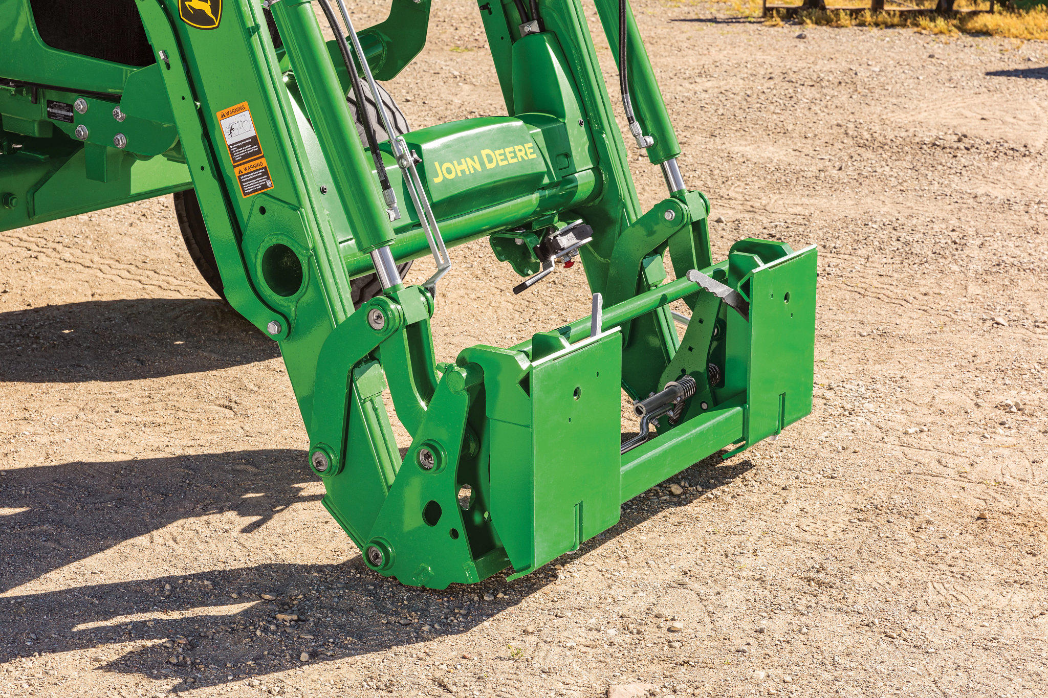 A close-up view of a green front loader attachment showing the hydraulic arms, hoses, and skid steer carrier adapter resting on a gravel surface