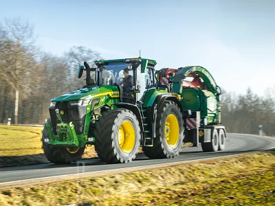 Tracteur 8R 540 roulant sur une route goudronnée en tractant un gros équipement vert, avec des champs d’herbe et des arbres en arrière-plan