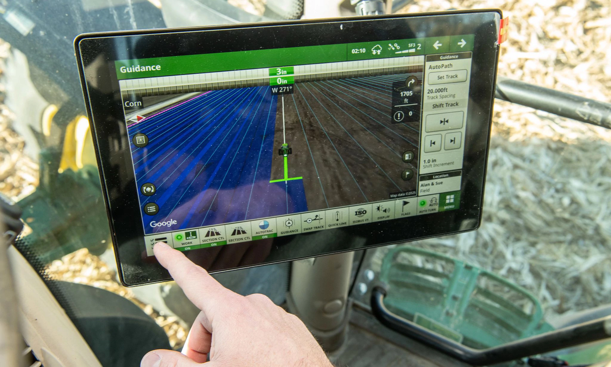 Close-up of John Deere in‑cab touchscreen display showing precision guidance lines and field view, with an operator’s hand pointing at the screen; mounted inside a tractor cab during field operation.