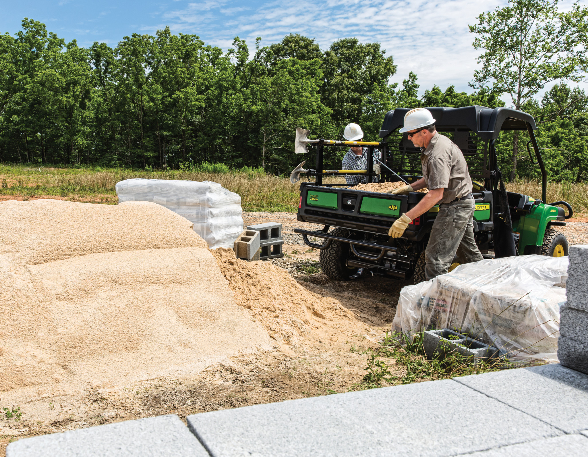Two white-helmeted landscapers working around an HPX615E Gator loaded with sand on a tree-lined jobsite.