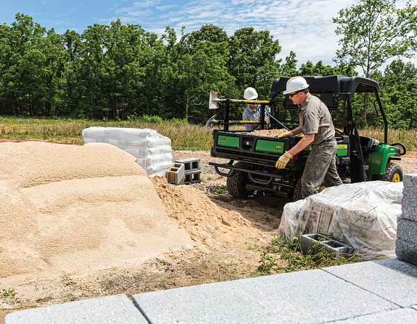 Two white-helmeted landscapers working around an HPX615E Gator loaded with sand on a tree-lined jobsite.