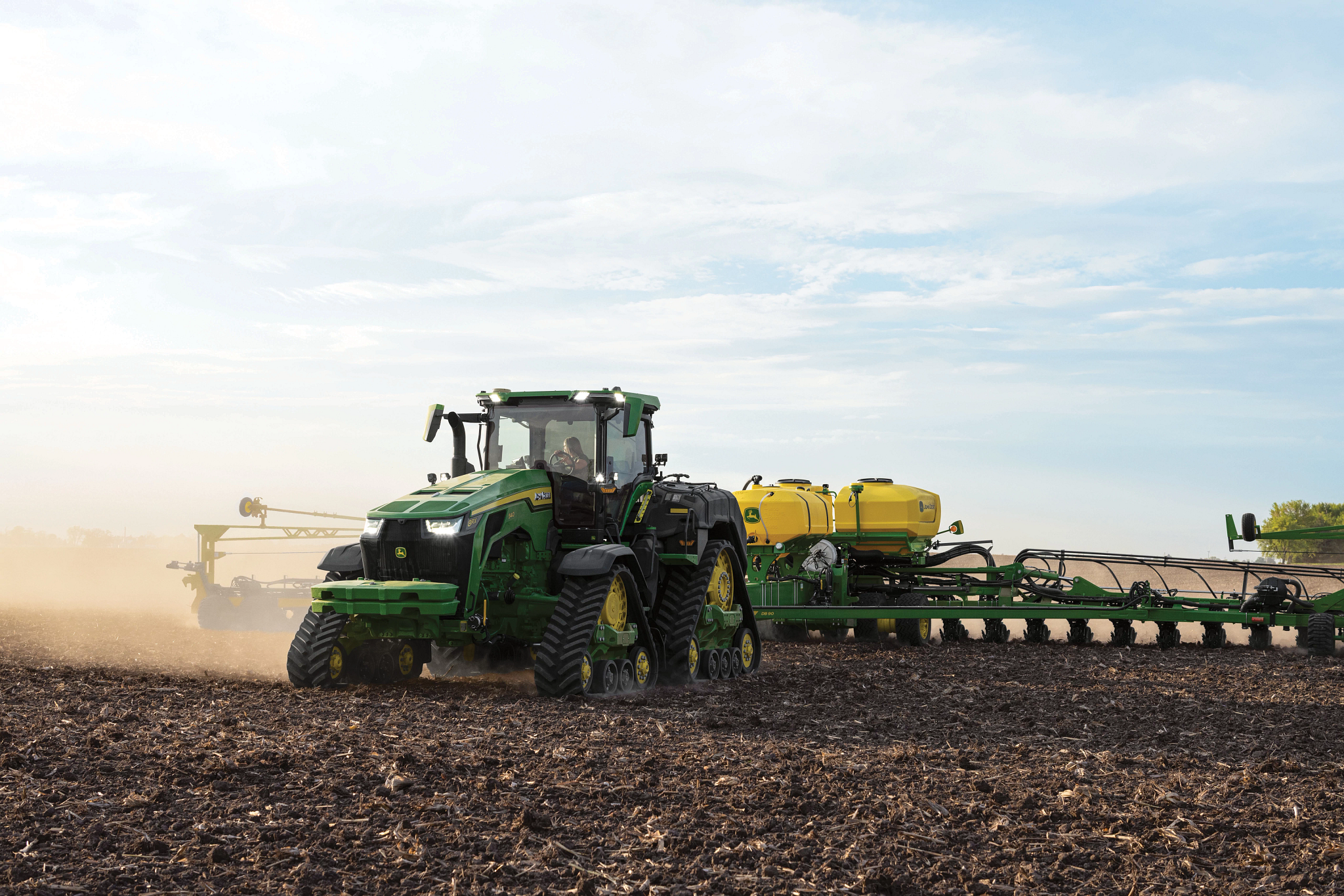 Green tractor pulling a wide planter across a dry, freshly tilled field. Dust rises behind the equipment as it moves under a lightly overcast sky, with multiple yellow seed tanks visible and open farmland stretching into the distance.