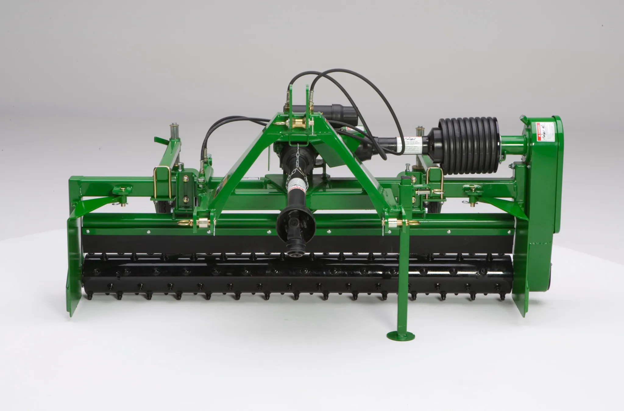 Front view of a rotary tiller or flail mower with PTO shaft, hydraulic hoses, and a wide drum with black blades, shown against a plain light background.