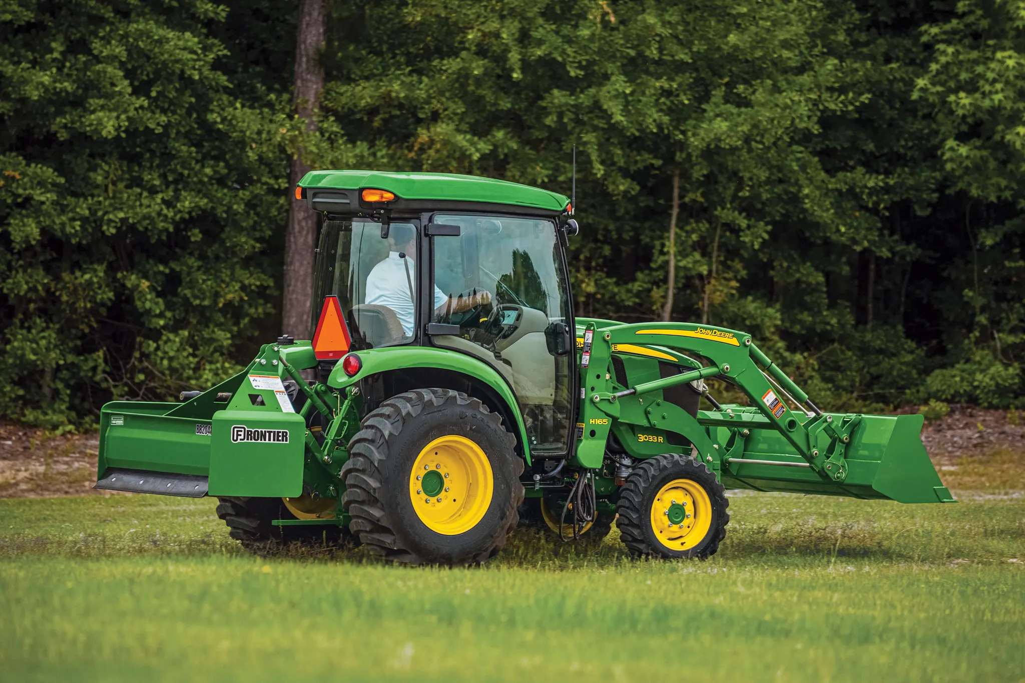 Side view of a green tractor with a front loader driving across a grassy field with an operator inside the enclosed cab and trees forming a dense wooded background