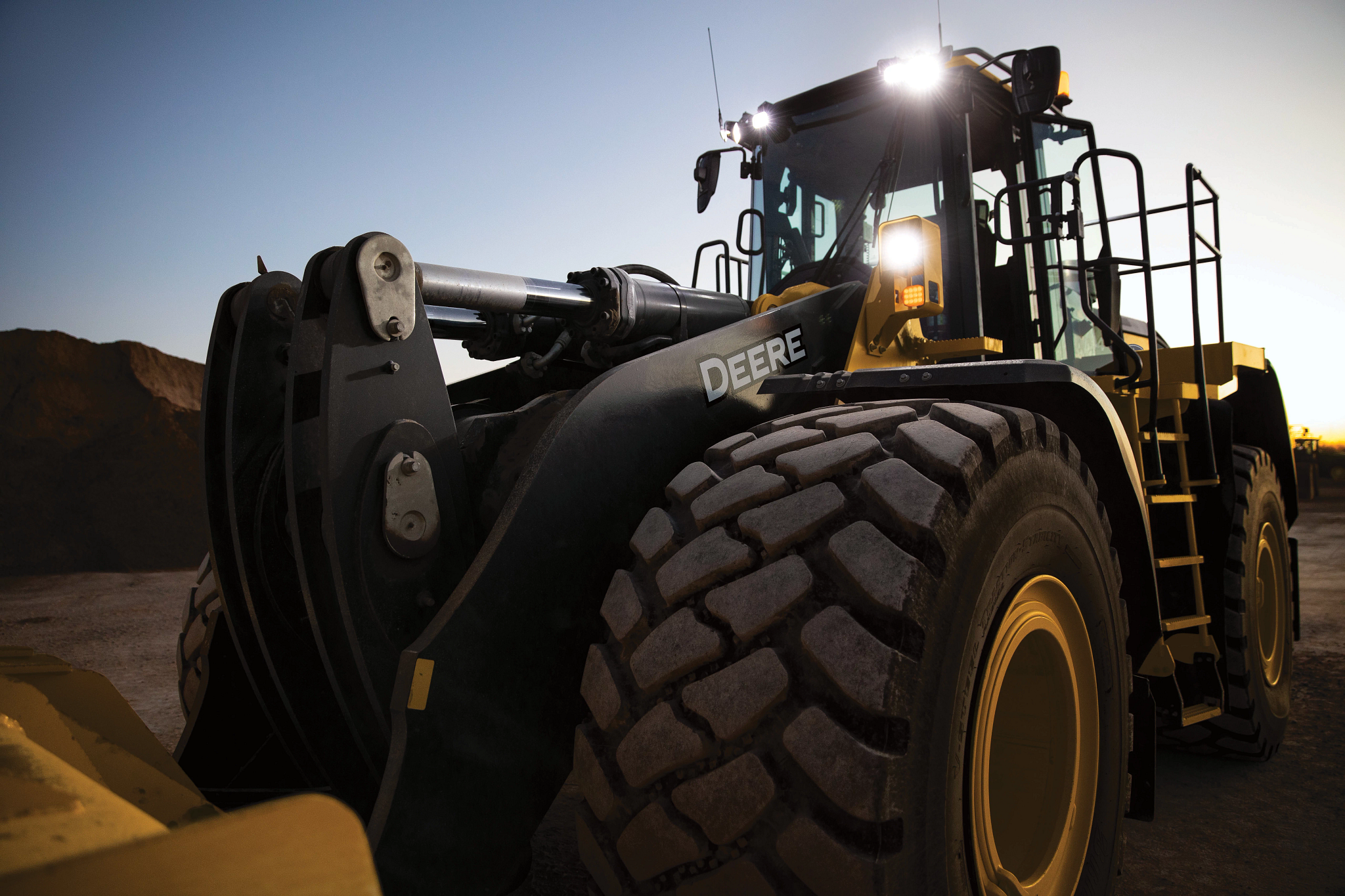 Large John Deere wheel loader with oversized treaded tires and a front bucket attachment parked at a work site, illuminated by bright headlights against a dim sky, with piles of dirt visible in the background