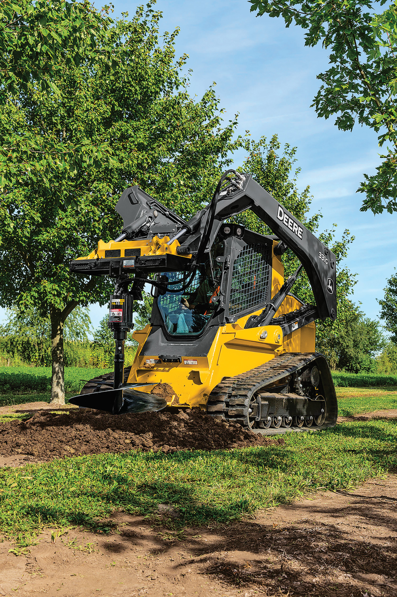 Front left view of compact track loader with front-mounted auger attachment drilling into dirt on a grassy area beneath trees