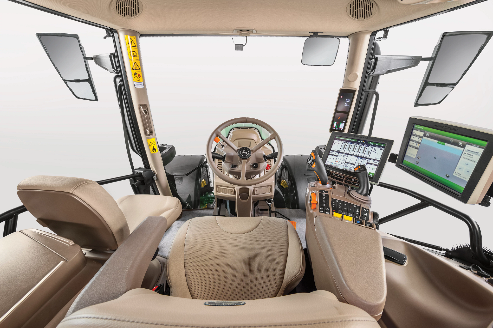 Interior view of John Deere tractor cab showing steering wheel, operator seat, armrest controls, touchscreen displays, joystick, mirrors, and overhead vents, photographed from behind the seat against a light background.