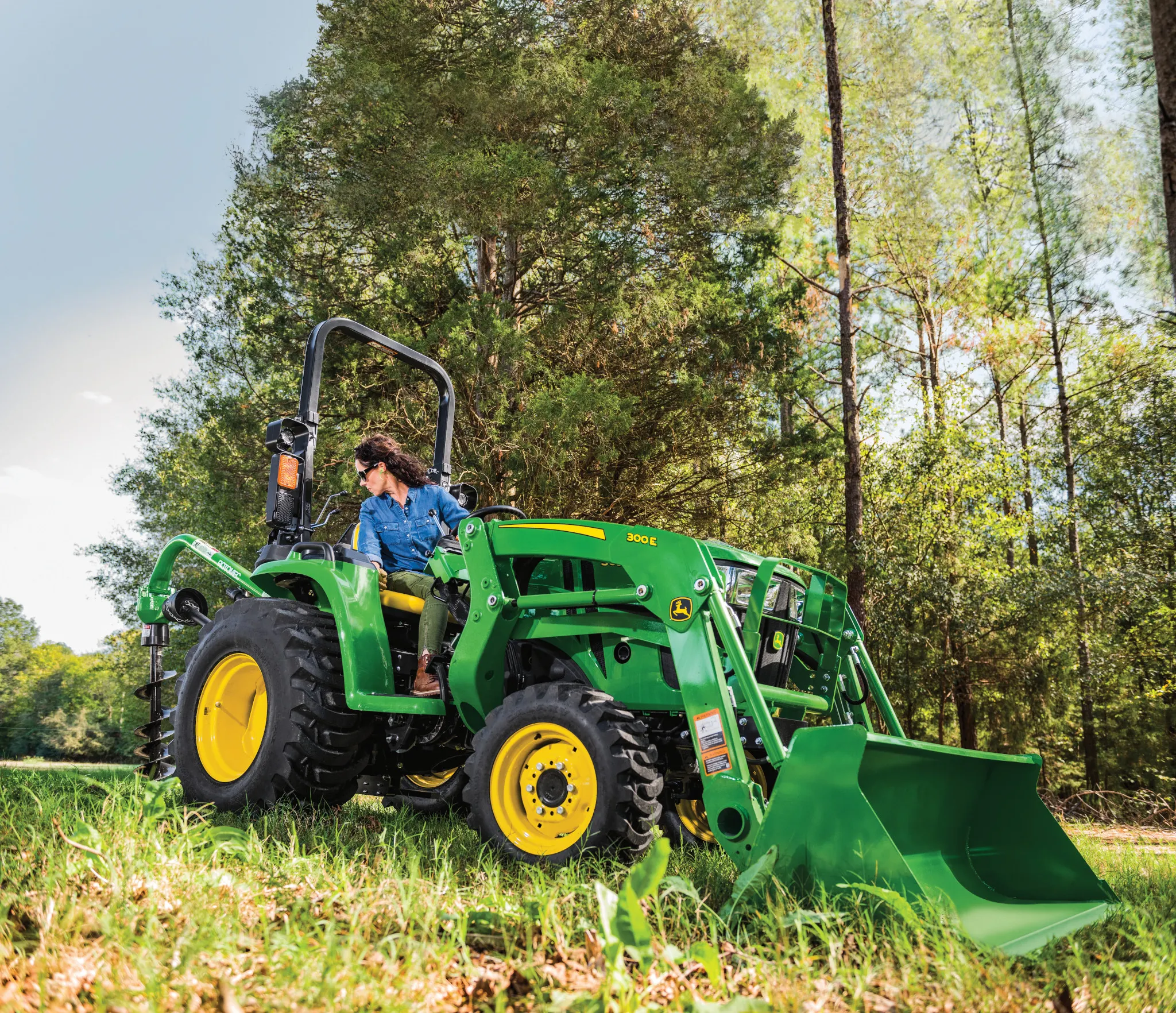 Person on a bright green tractor with a front loader attachment using a rear-mounted auger in a grassy field, with tall trees in the background under partly sunny conditions