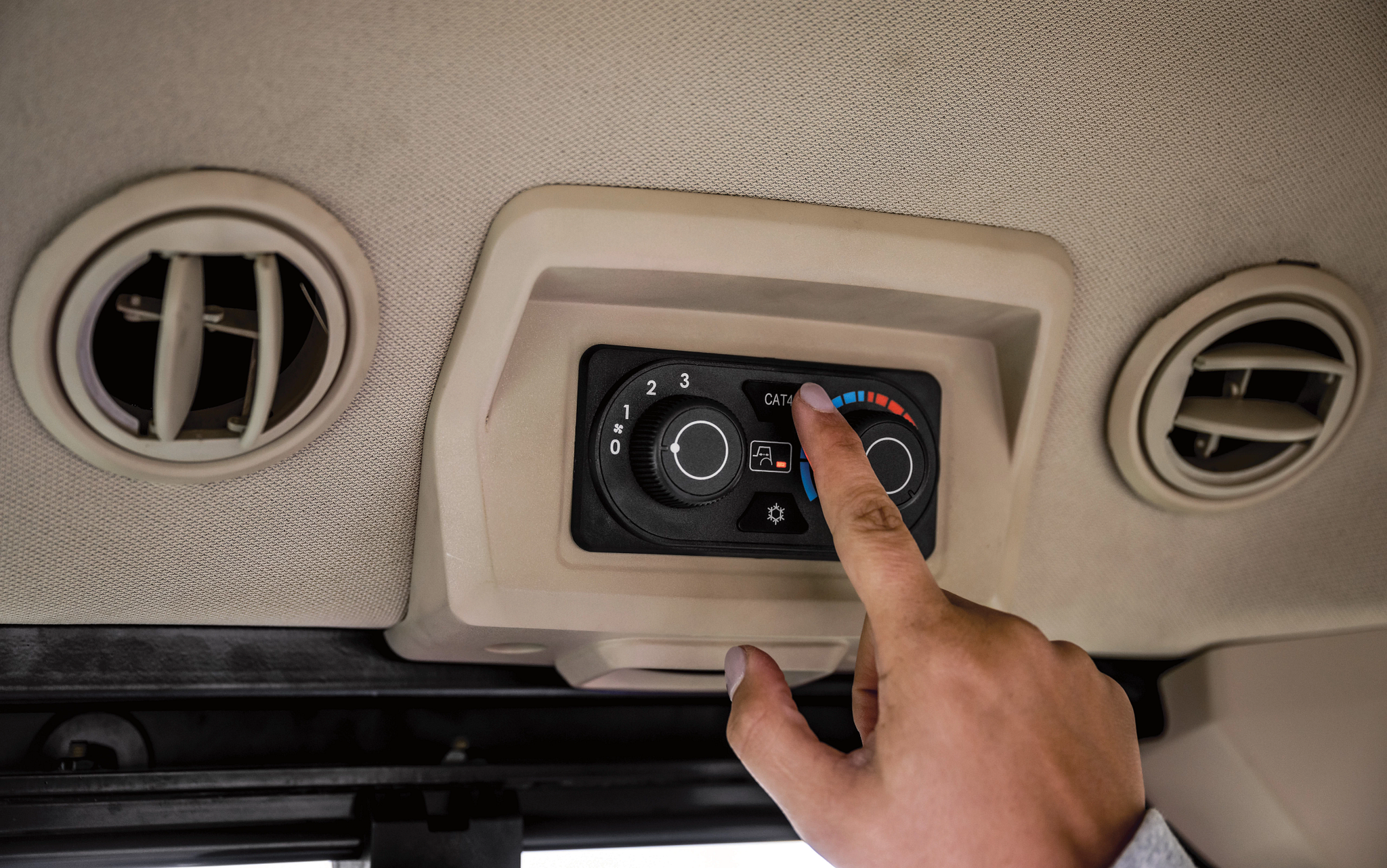 Hand pressing a button on an overhead climate control panel inside a John Deere equipment cab, with round air vents on both sides and illuminated temperature and fan icons on the control display.