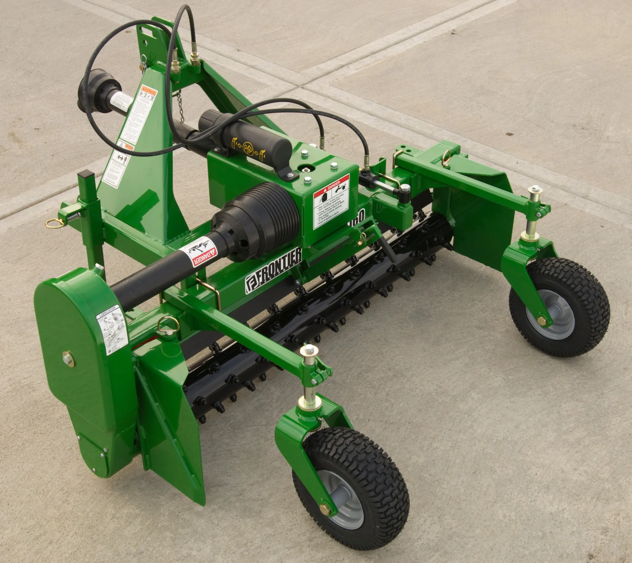 Green agricultural machine with a steel frame, hydraulic motor and hoses, rotating drum, and four rubber wheels, shown outdoors on a concrete surface with warning labels and manufacturer branding visible.