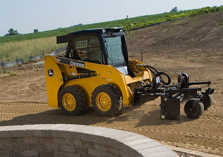 Side right view of a 315 skid steer using a power rake front attachment on a dirt lot next to a brick retaining wall