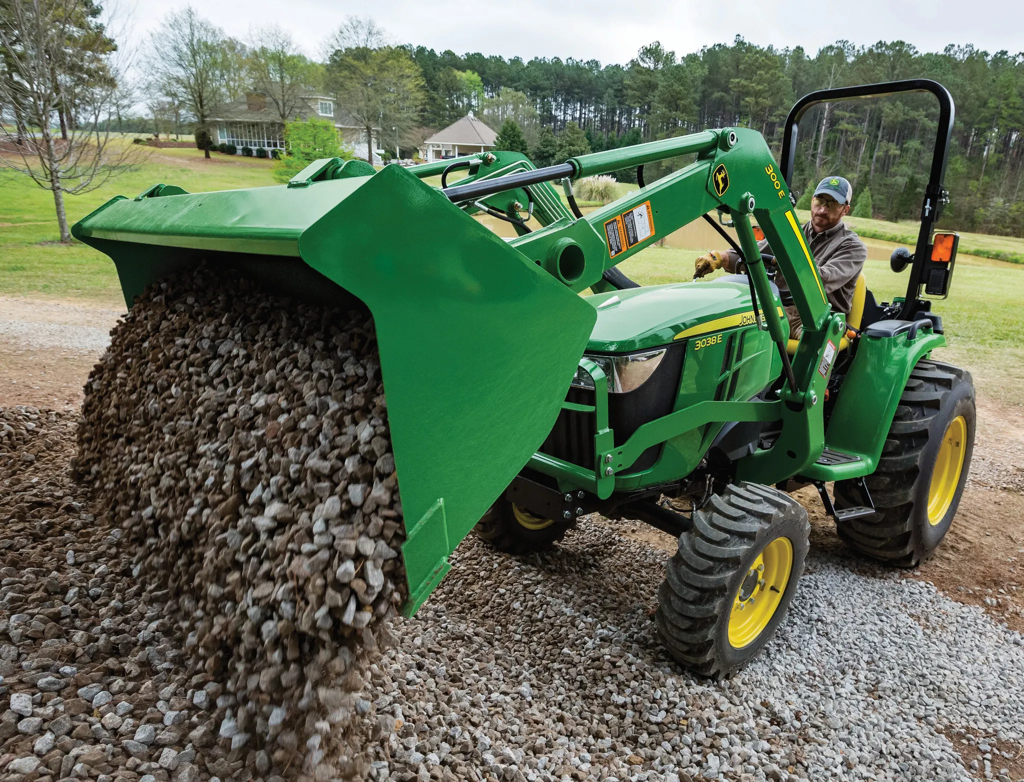 Green tractor with a front loader tipped forward, pouring a large load of gravel onto the ground and set in an open yard with grass, trees, and buildings visible in the background