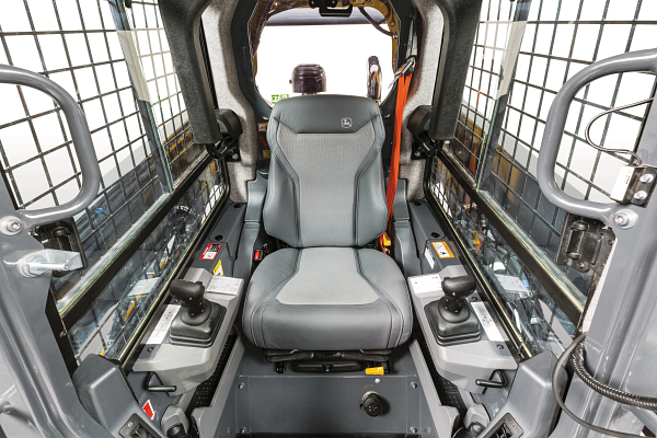 Interior cab view of a skid steer loader showing a central padded operator seat, protective side grid panels, and dual joystick controls on both armrests