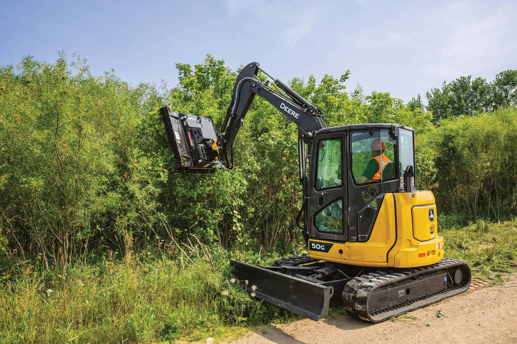 Compact track excavator with a brush cutter trimming dense vegetation along the edge of a path