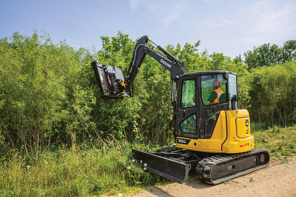 Compact track excavator with a brush cutter trimming dense vegetation along the edge of a path