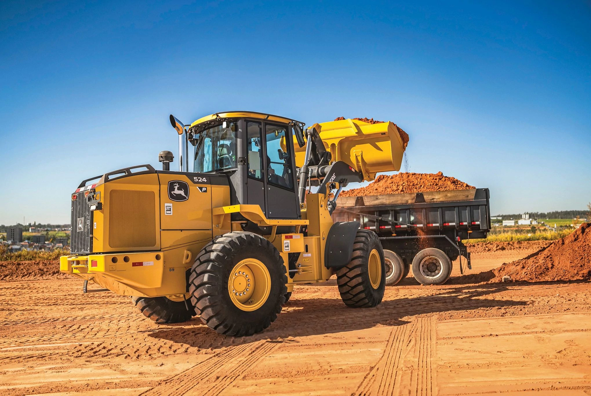 A 524 P-Tier Wheel Loader scoops reddish-brown dirt into the bed of a black dump truck at a construction site.