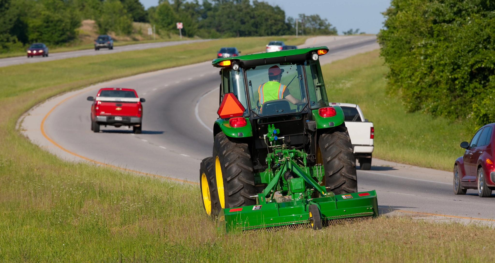 Rear view of a tractor with a rear-mounted mower cutting grass along the shoulder of a roadway while cars drive past on a curved two‑lane road