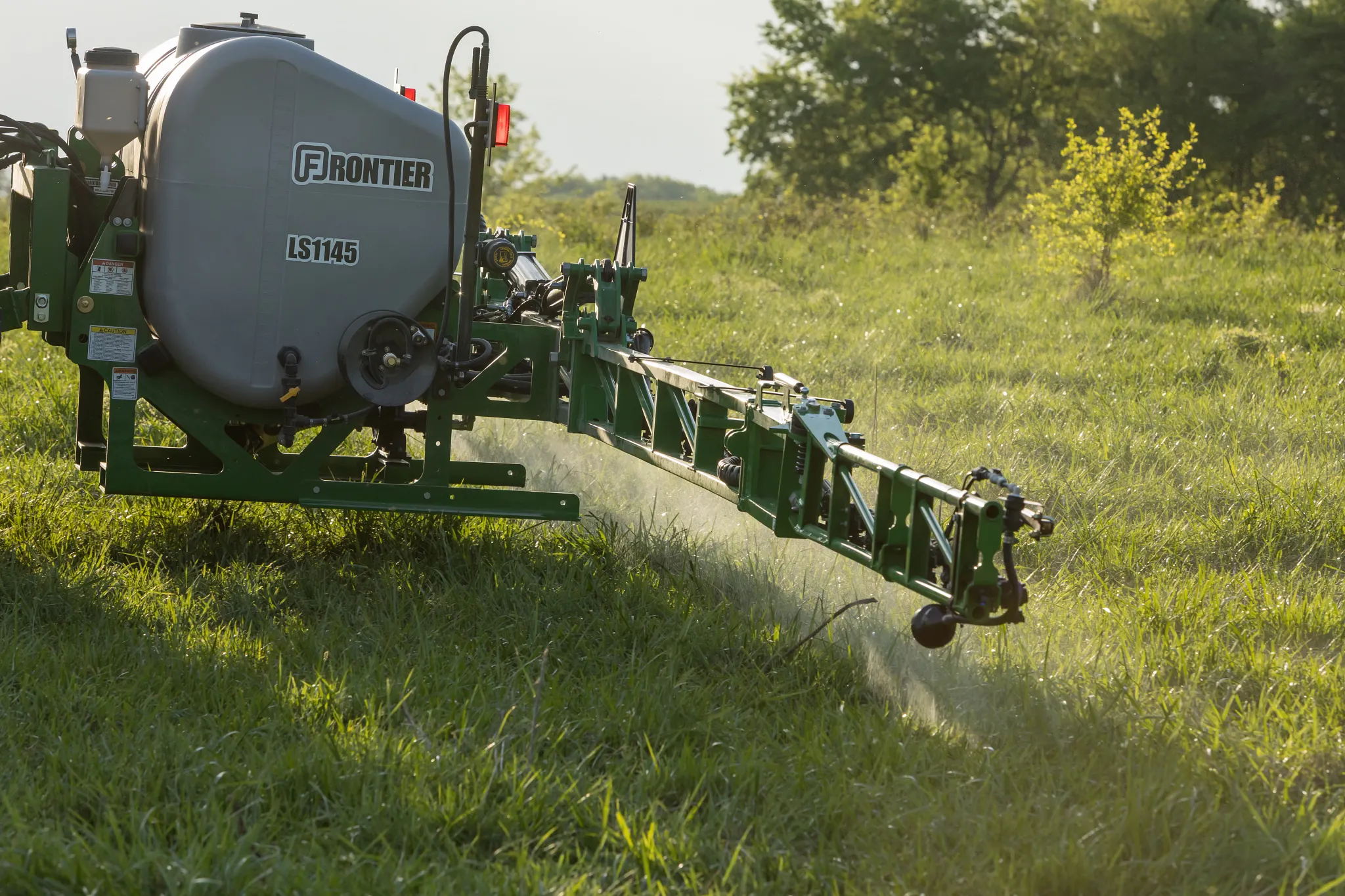 Rear-mounted agricultural sprayer labeled Frontier LS1145 with boom extended, spraying liquid over tall grass in a sunny field with trees in the background.