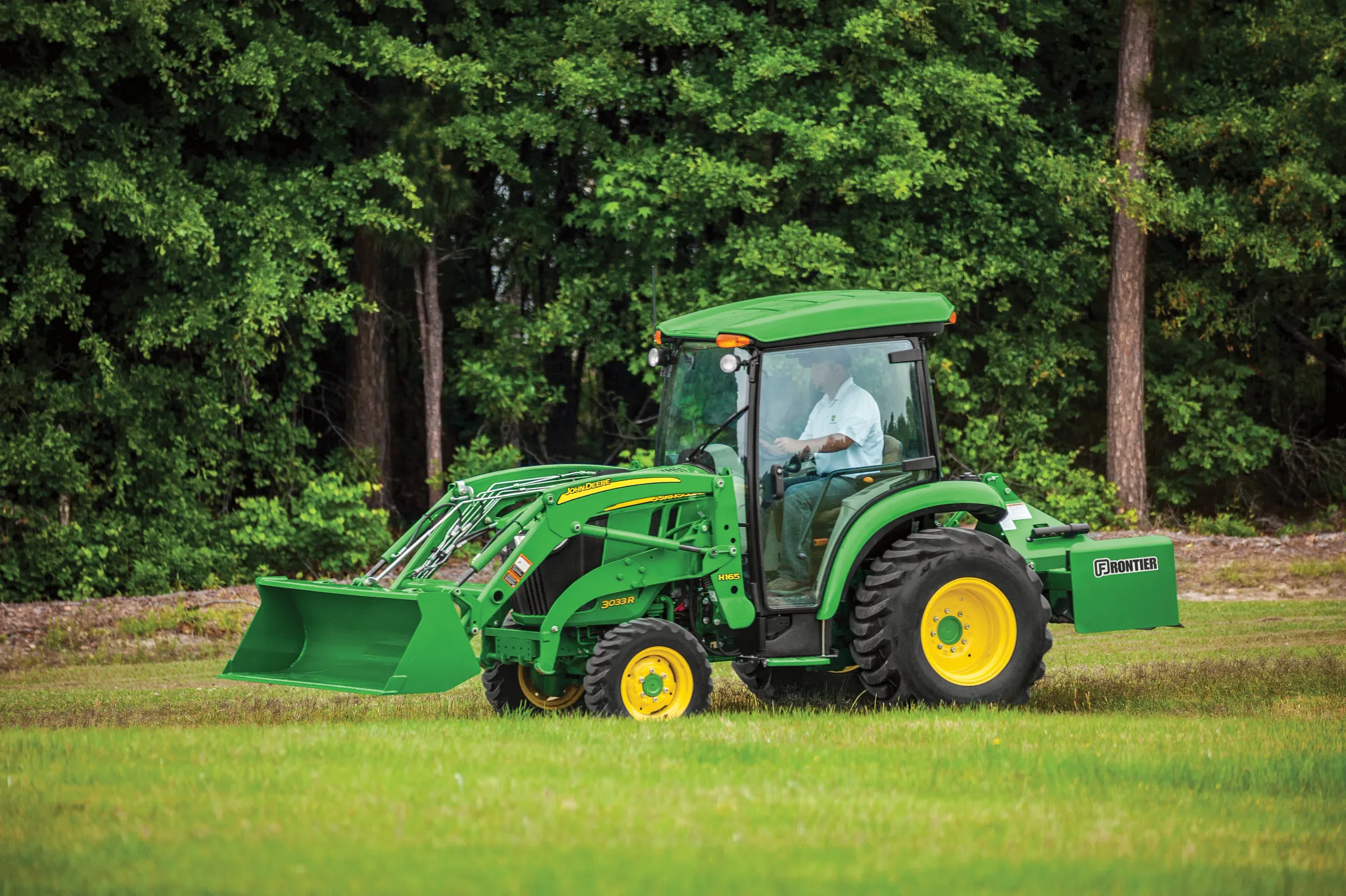 Green tractor with yellow wheels driving across a grassy field, equipped with a front loader bucket and enclosed cab with trees forming a dense backdrop