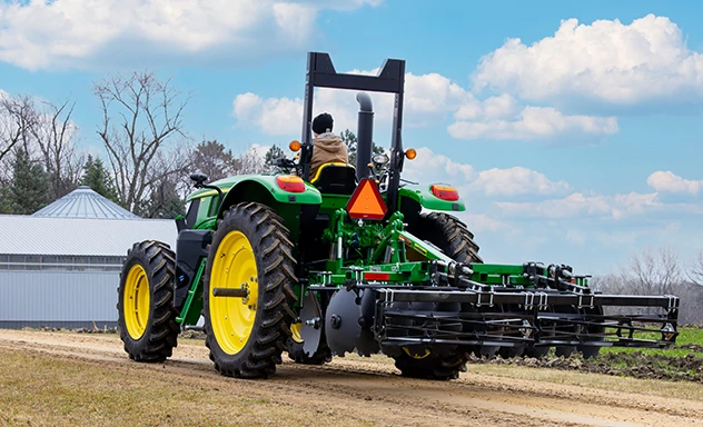 Rear view of a 6M Tractor with a TM13 Series Frontier Tandem Disk Harrow driving through a farmyard.