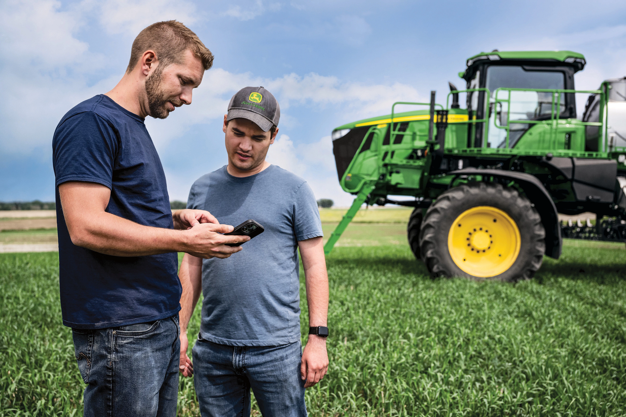 Two people standing in a green crop field look at a smartphone together, while a large green agricultural sprayer with yellow wheels is parked nearby under a partly cloudy sky