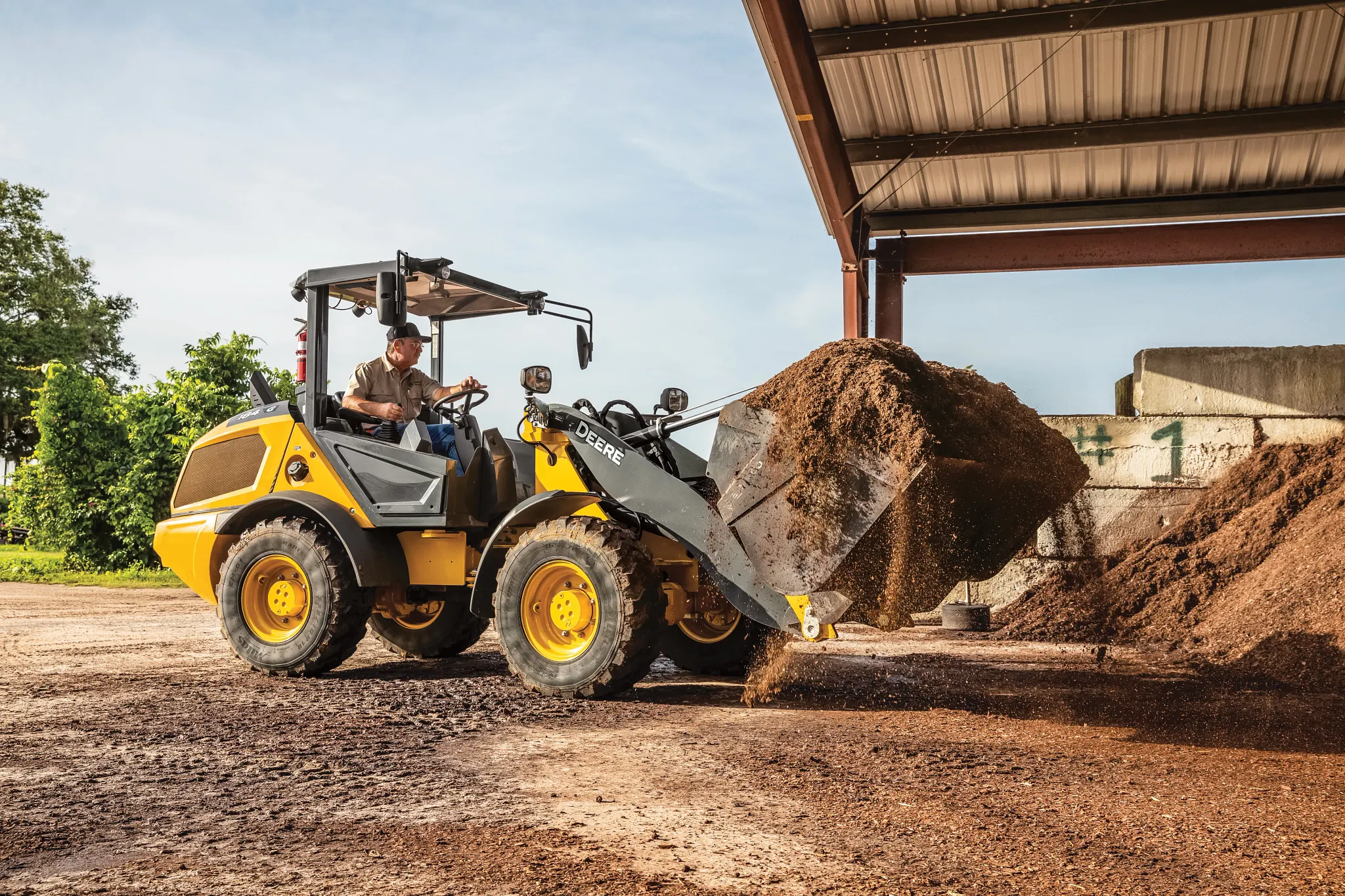 A wheel loader lifting a bucket of loose soil inside an open-sided structure, with material piles on the ground and daylight visible beyond the roofed area.